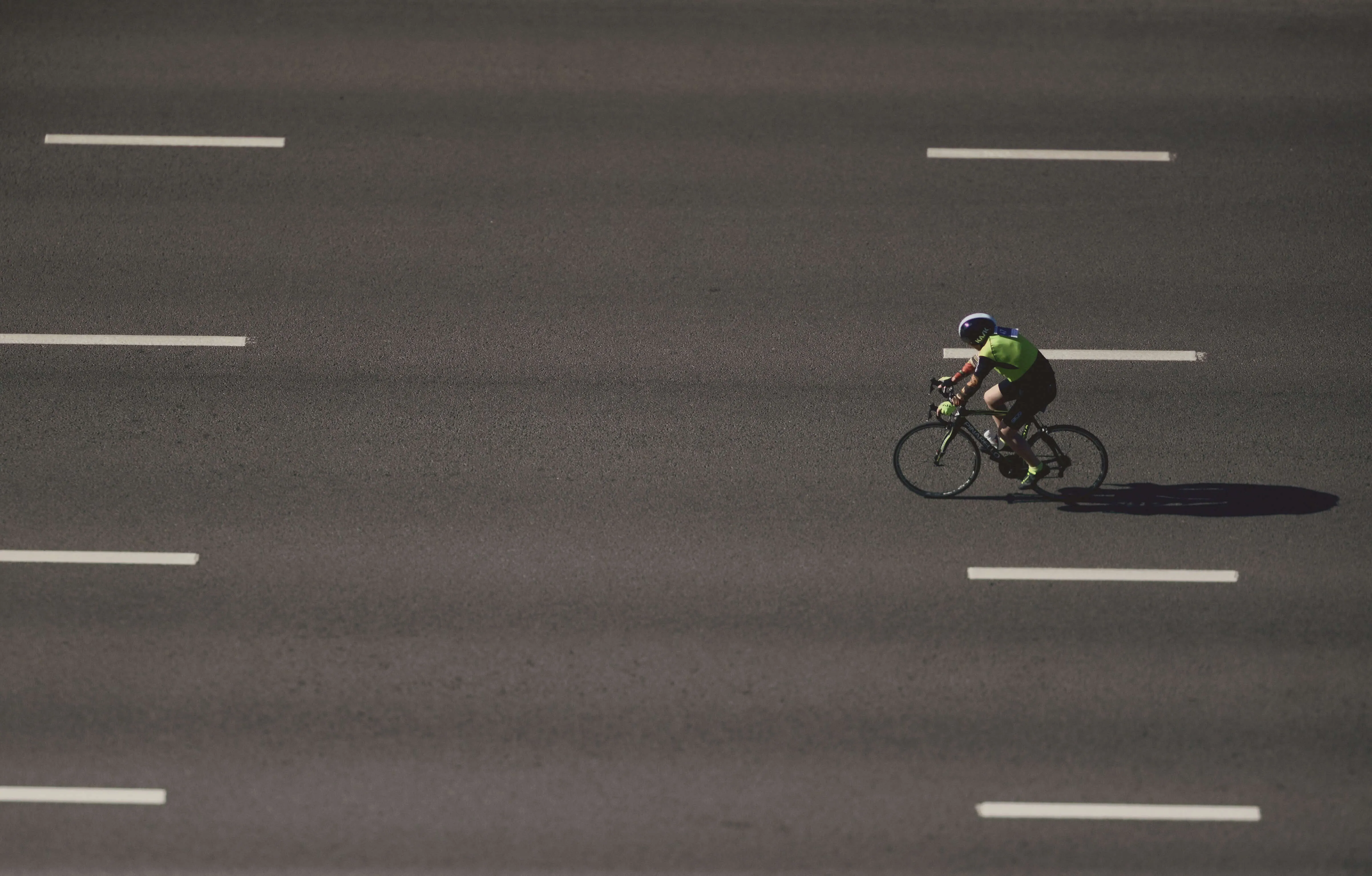 Aerial view of a cyclist in bright green athletic wear riding across a dark asphalt surface marked with white lane dividers, casting a long shadow while moving through the geometric pattern of road markings, symbolizing choice, determination, and purposeful movement forward